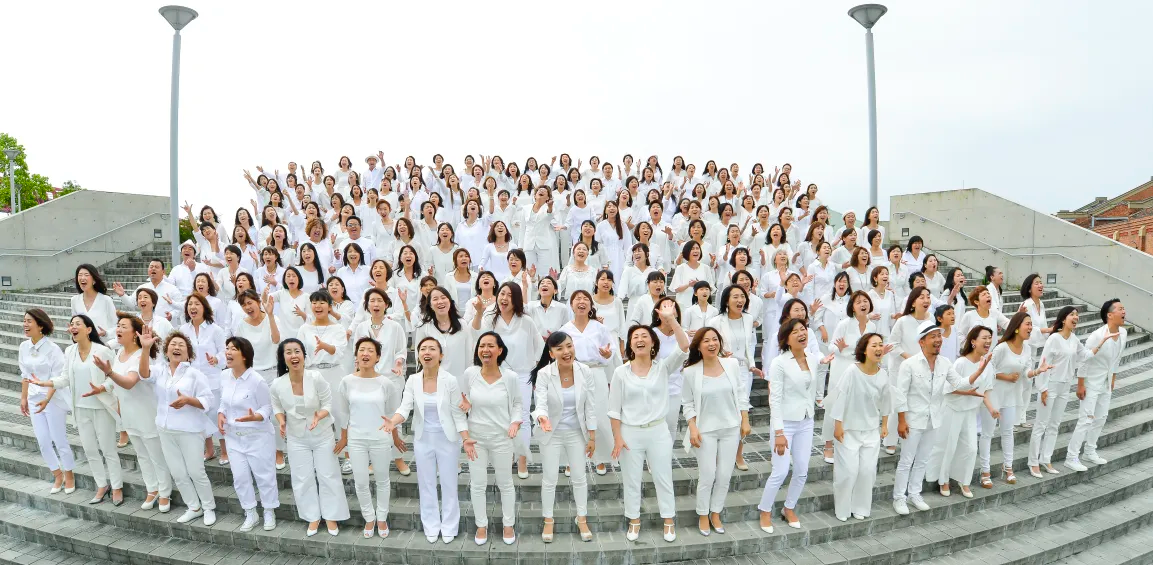 階段状の屋外ステージで、白い衣装を着た大人数のシンガーグループが笑顔で歌っている写真